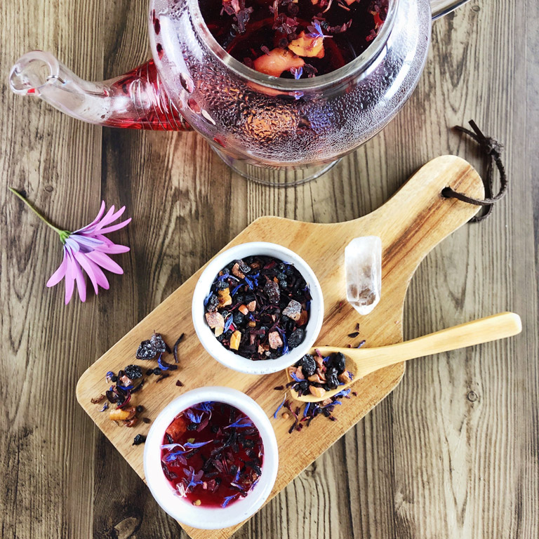 A glass teapot and two small teacups sit on a wooden table filled with steeping Blue Moon tea.