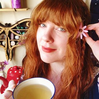 Photo of Sarah Louise, a woman with blue eyes and red hair holding a teacup and purple flower. In the background are a red teapot with white spots, a small vase of purple flowers, and a crystal display shelf in the shape of a luna moth.