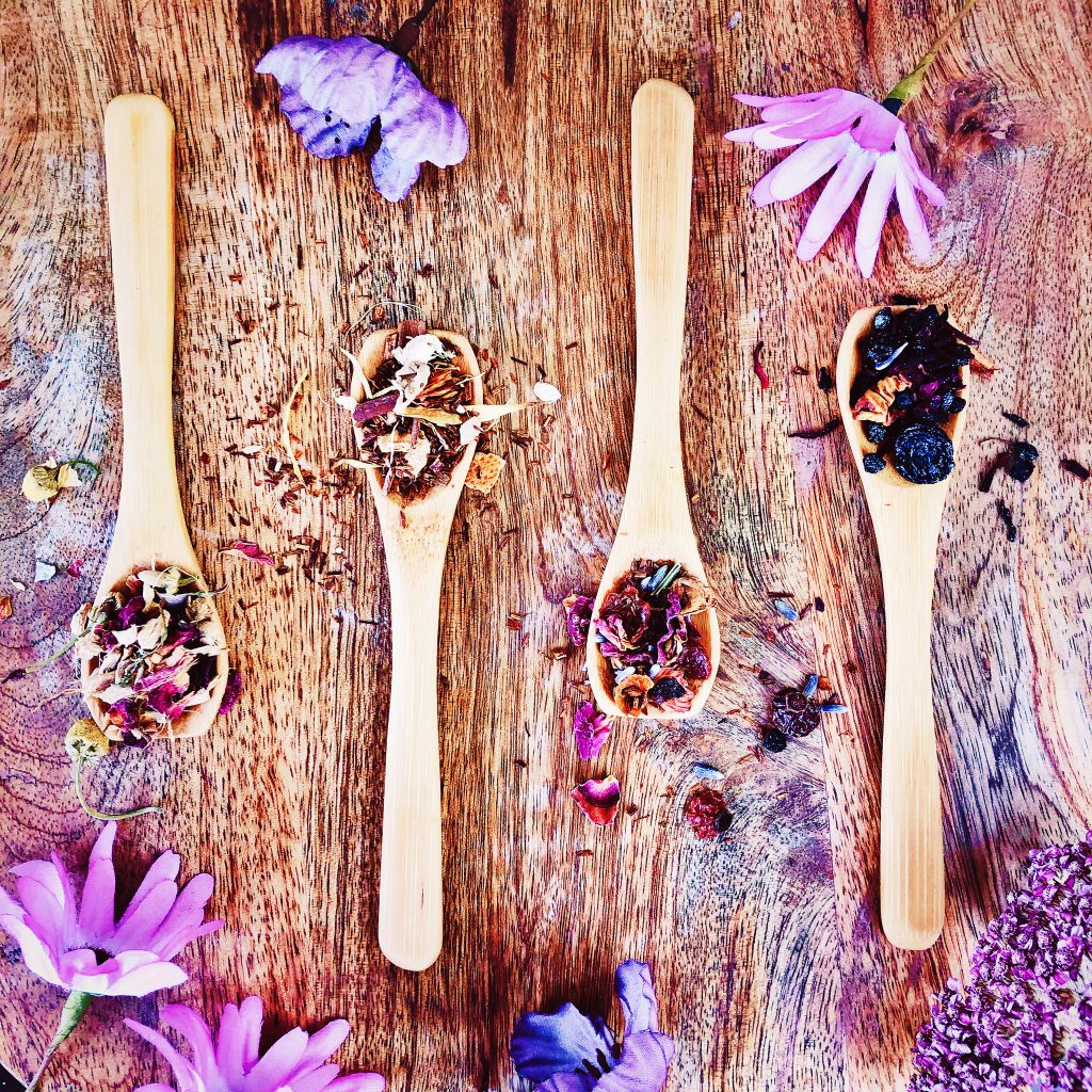 Four teaspoons heaped with the four different tea blends included in this bundle. From left to right: Whispered Wishes herbal blend, Ritual At Dawn rooibos and floral blend, Ritual At Dusk rooibos and spices blend, and Blue Moon berry and floral blend.